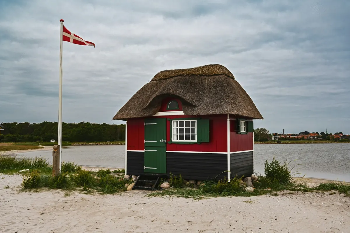 Lite stråtakhus i rødt og grønt på en sandstrand med dansk flagg
