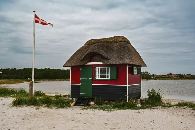 Lite stråtakhus i rødt og grønt på en sandstrand med dansk flagg