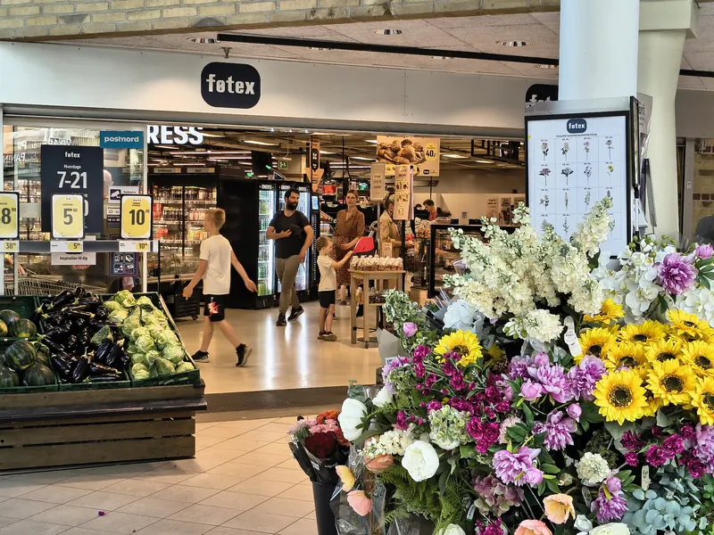 Inngangen til en dansk dagligvarebutikk med grønnsaker, blomster og prisskilt i kroner