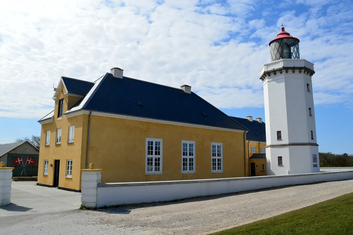 Hvitt fyrtårn ved siden av et gult fyrmesterhus under blå himmel