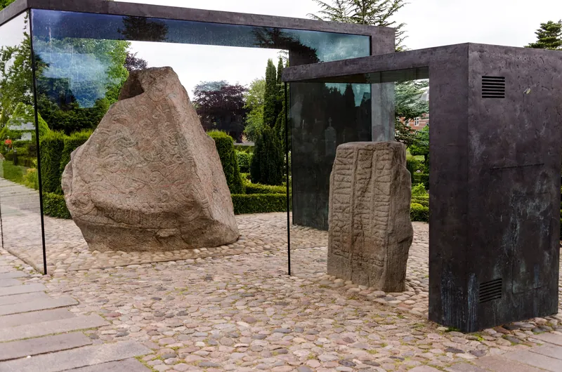 De to runesteinene i Jelling i beskyttende glassmontre på brosteinsplassen ved kirkegården