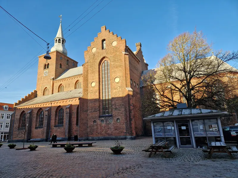 Gammel teglsteinskirke med trappegavl og tårn ved et torg i Randers i høstsol