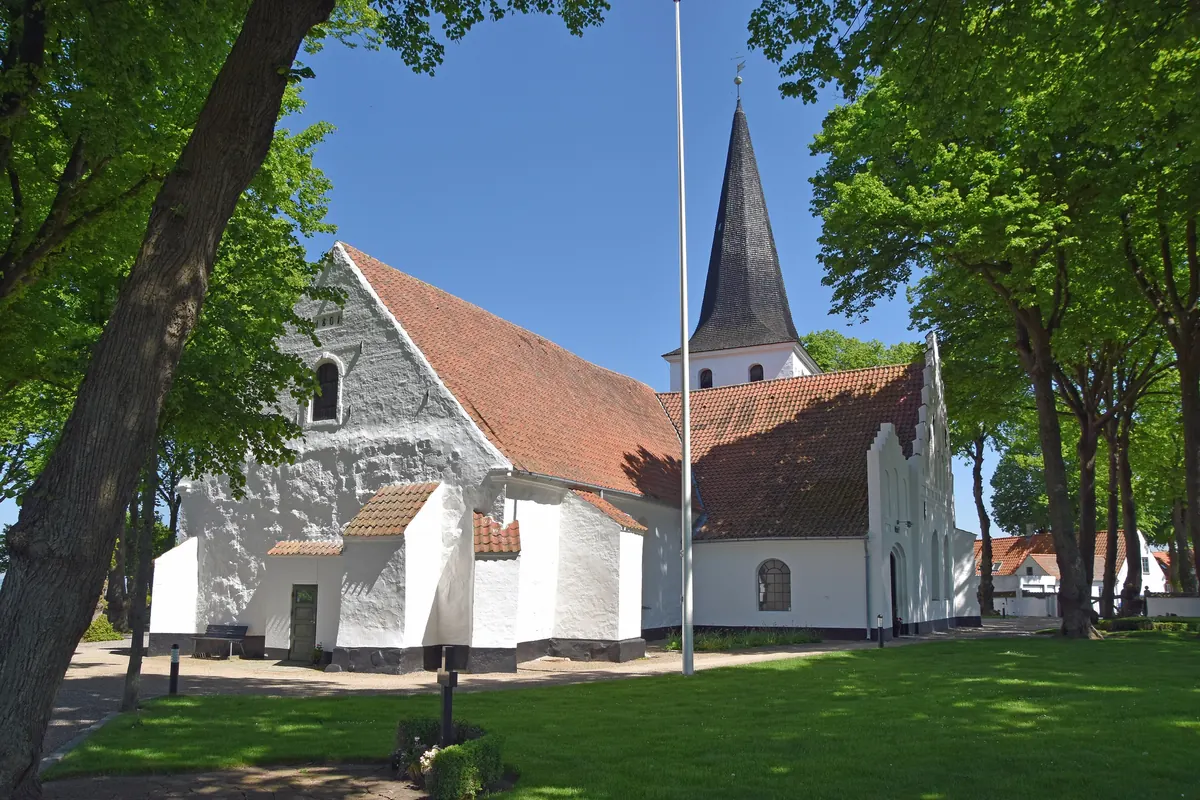 Hvitkalket kirke med spisst tårn mellom grønne trær i Bogense