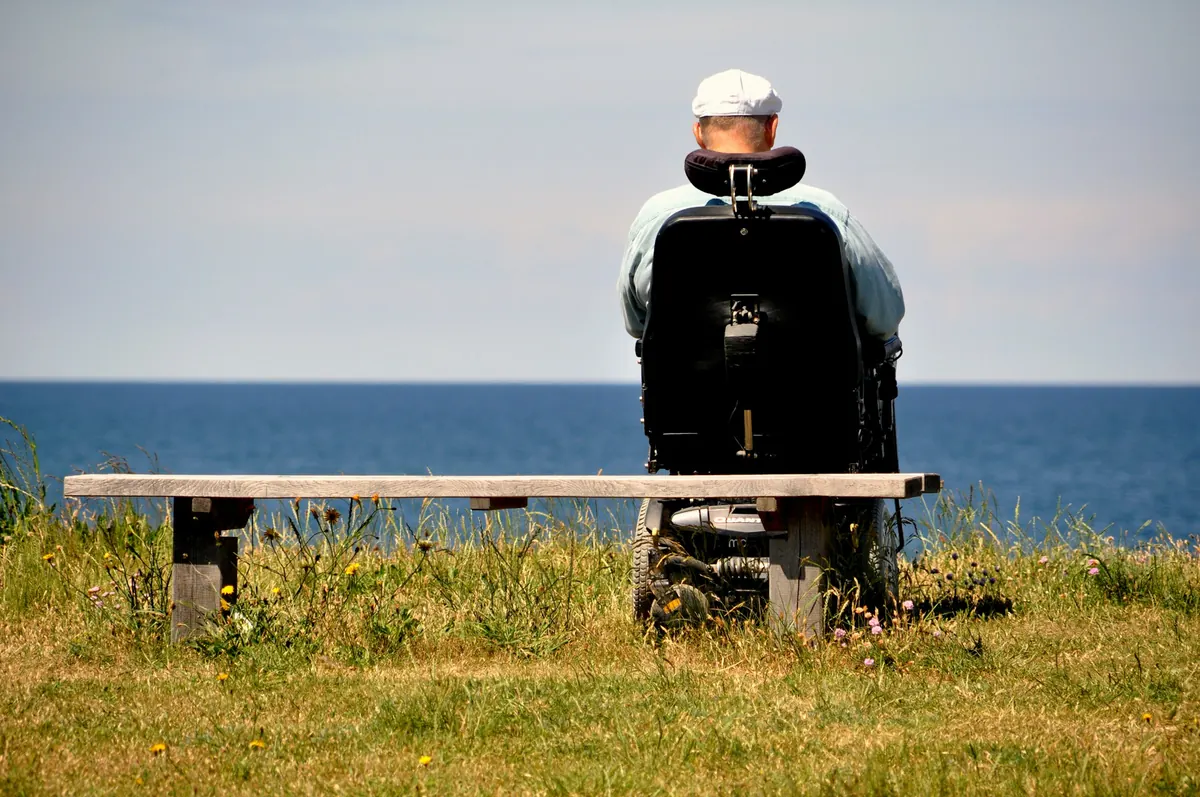 Person i elektrisk rullestol sett bakfra på en gressplen ved havet