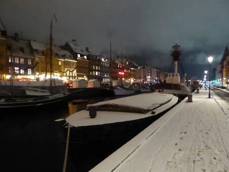Snødekt kai langs en kanal med gamle skip og opplyste hus en vinterkveld
