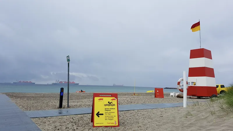 Livredderhytte med rødt og hvitt tårn og gult informasjonsskilt på en sandstrand en overskyet sommerdag