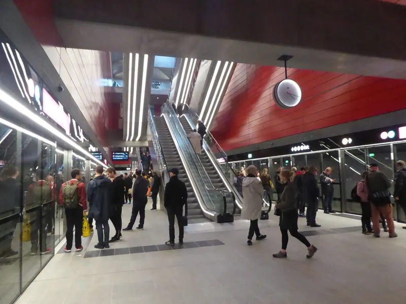 Reisende på perrongen på metrostasjonen ved København Hovedbanegård, med røde vegger og rulletrapper