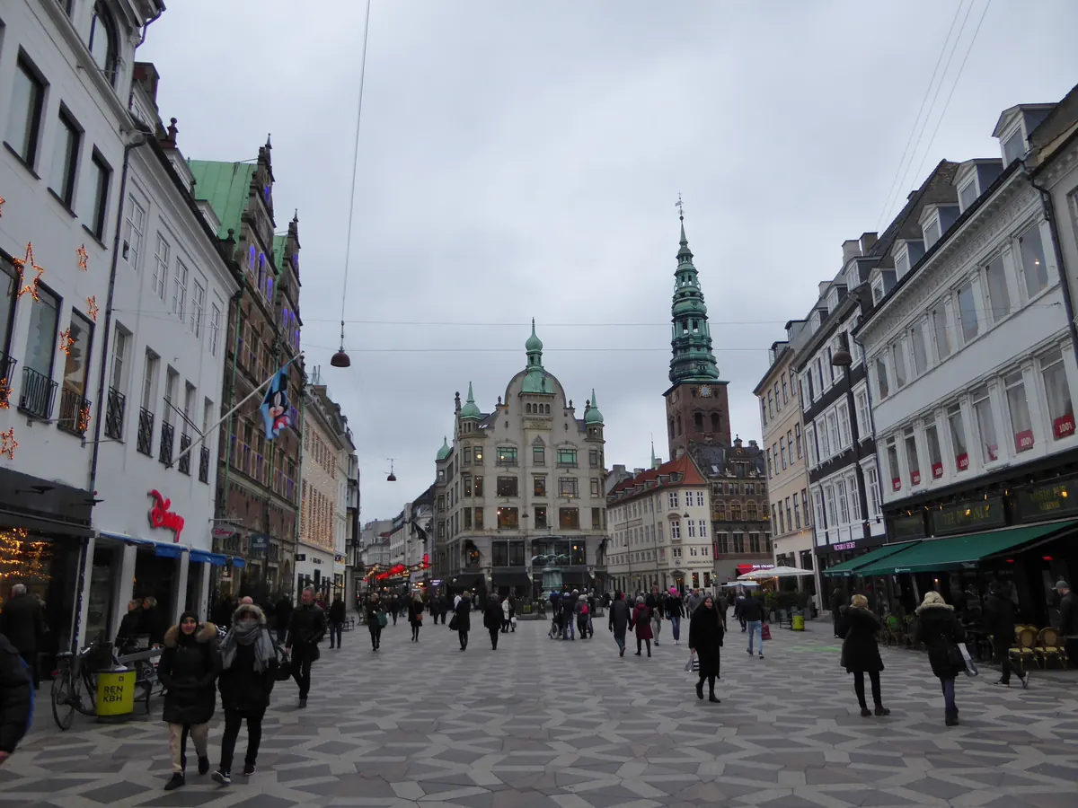 Gågaten ved Amagertorv i København med butikker og folk, og Nikolaj kirkes grønne spir i bakgrunnen