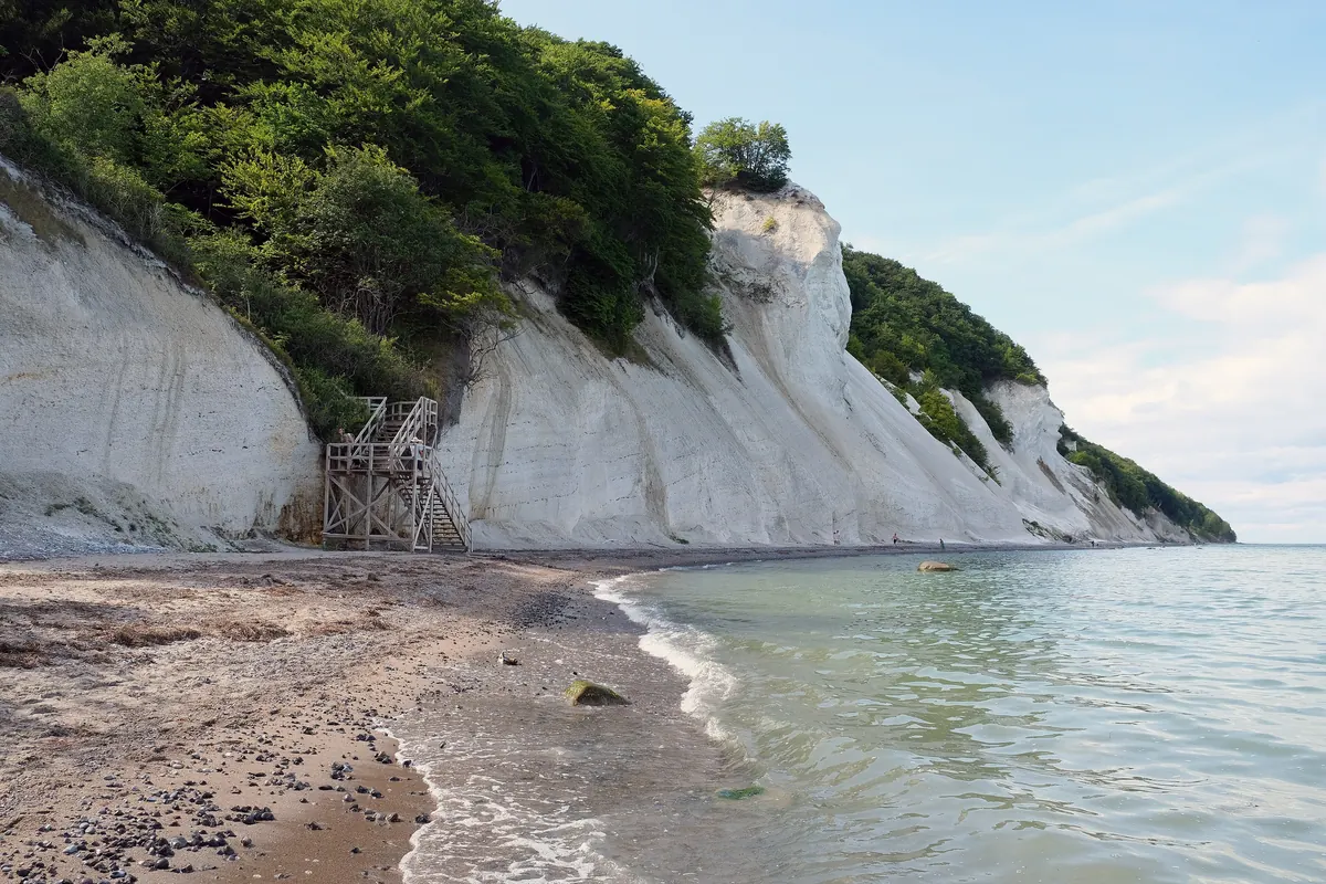 Hvite krittklipper med skog på toppen over en smal strand og klart, grønt vann