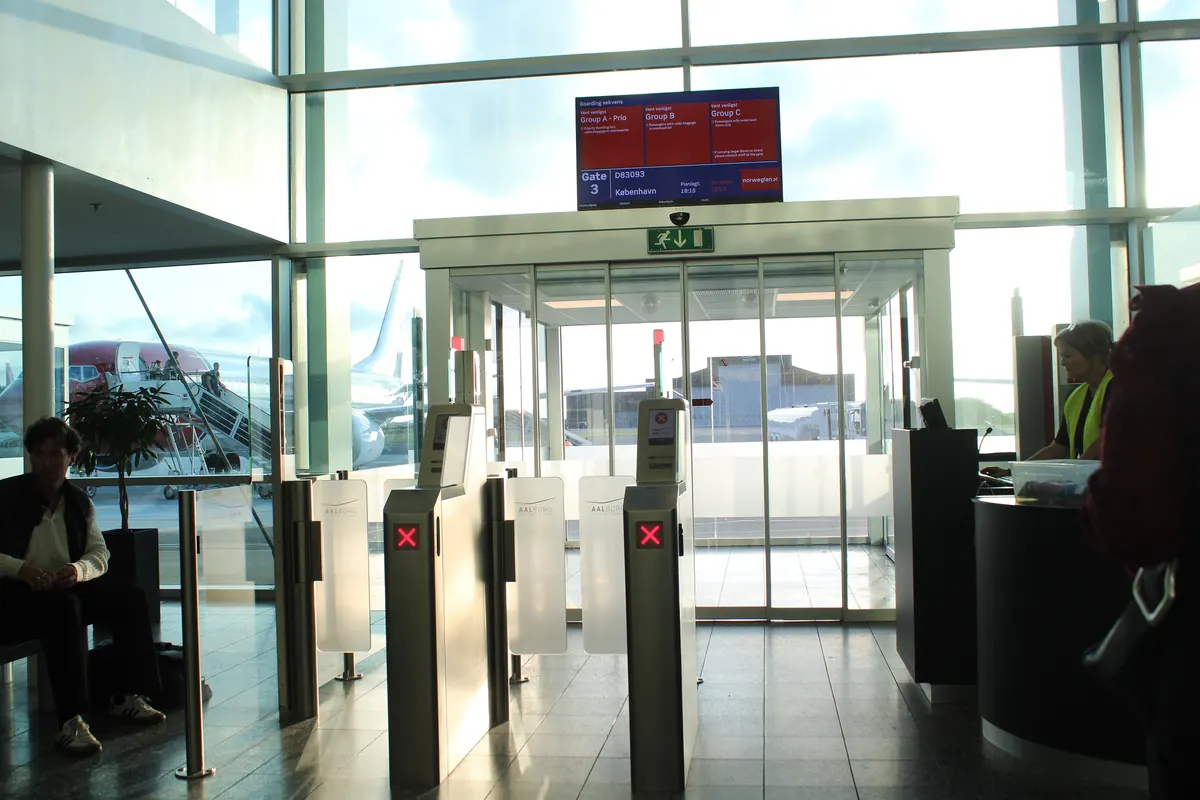 Sikkerhetsporter og avgangsskjerm i den lyse glassterminalen på Aalborg lufthavn