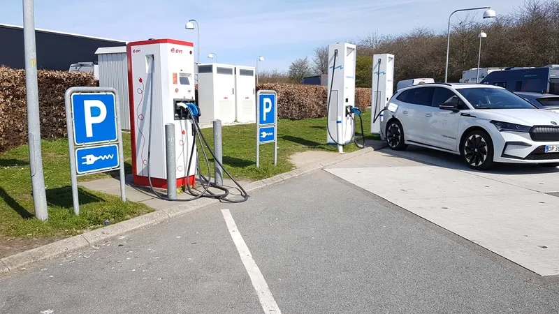 Hurtigladere og en hvit elbil ved en rasteplass med blå parkeringsskilt for lading