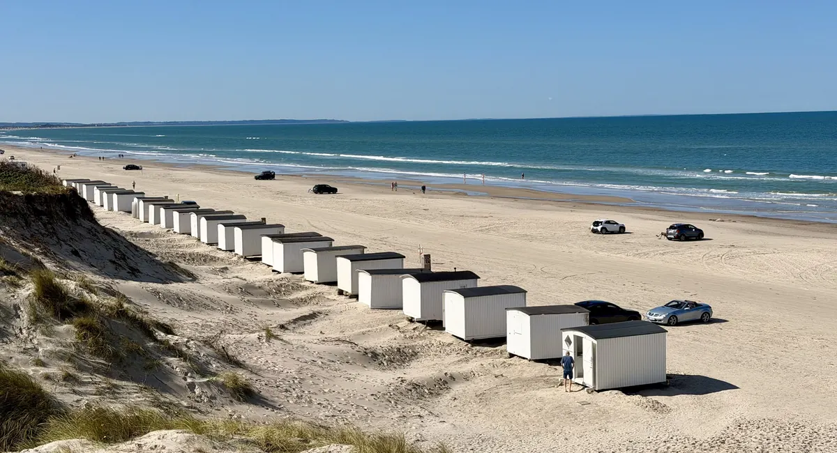 Rekke med hvite badehus og biler på den brede sandstranden ved Blokhus