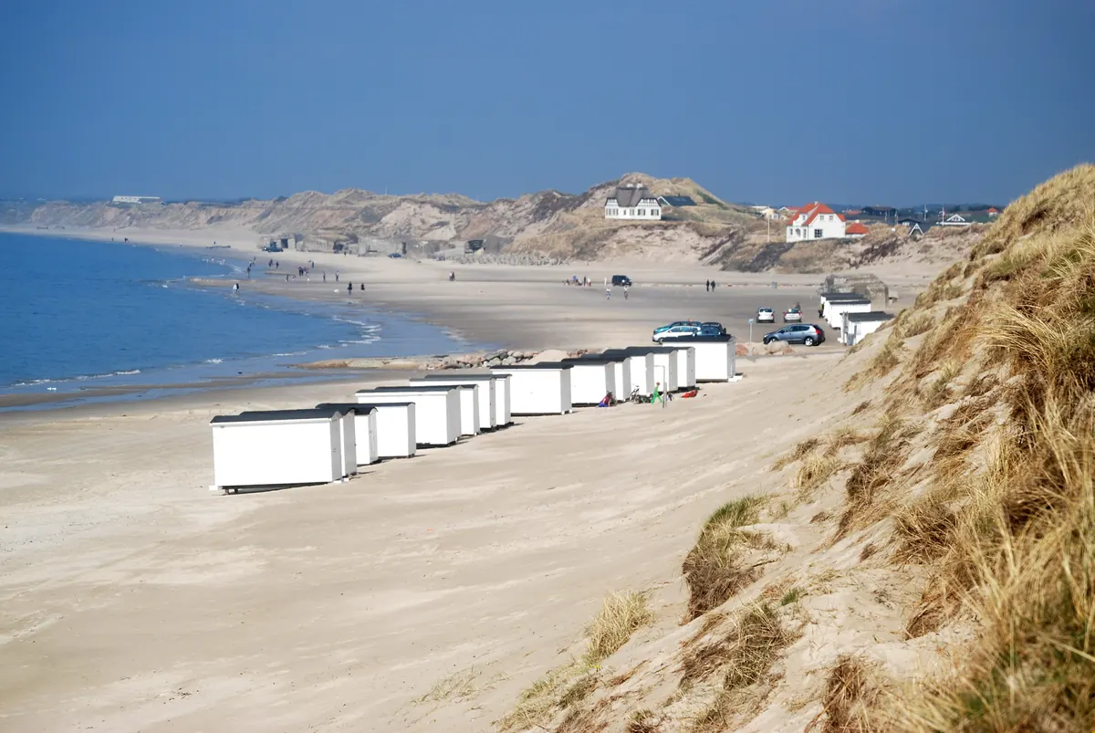 Rekke med hvite badehus på sandstranden ved Løkken, med klitter og hav i bakgrunnen