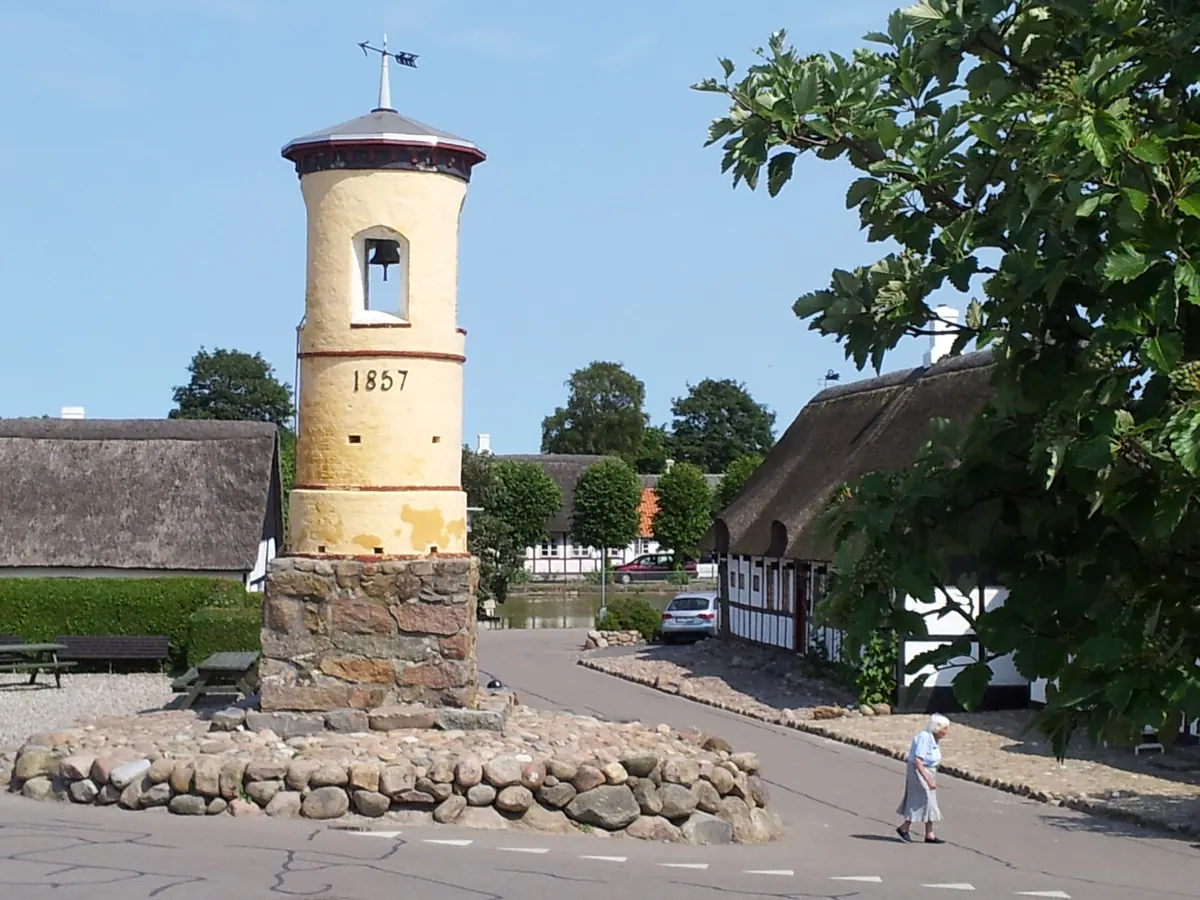 Gult klokketårn på en steinsokkel mellom stråtakshus i landsbyen Nordby på Samsø