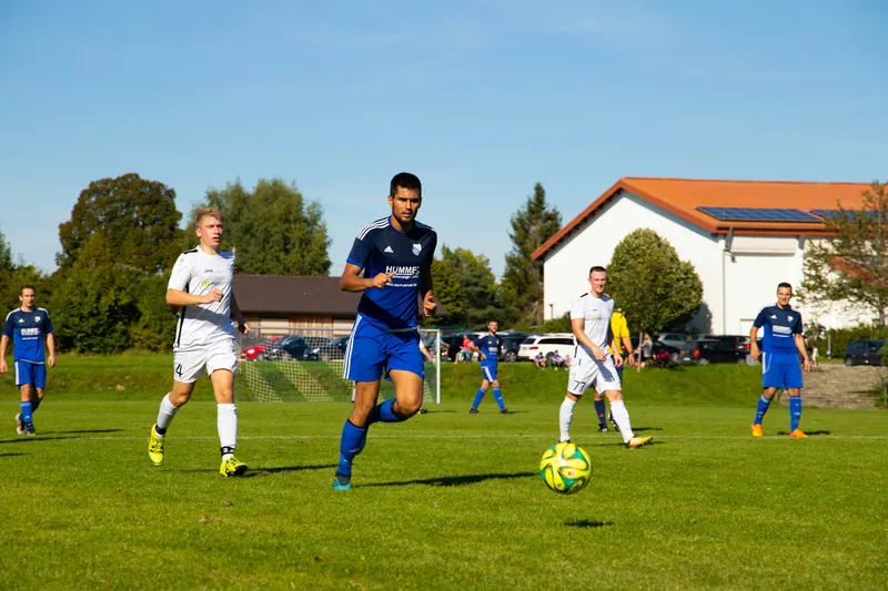 Fotballkamp på en gressbane med hus og trær i bakgrunnen en solrik dag